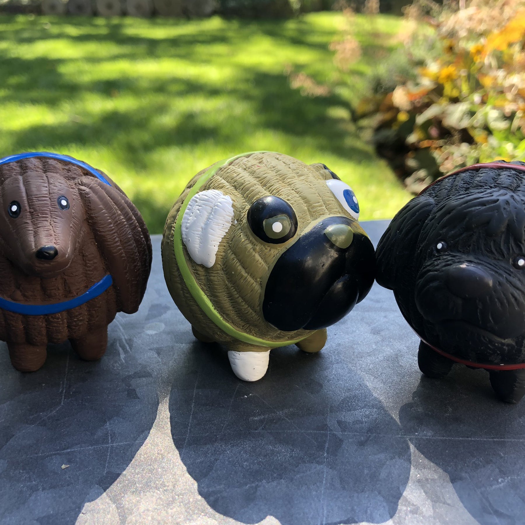 Lifestyle image of Finbar, Wally, and Puggie the dogs sitting next to one another. They're all outside sitting on a grey slab. WIth green grass in the background, bushes, and leaves.