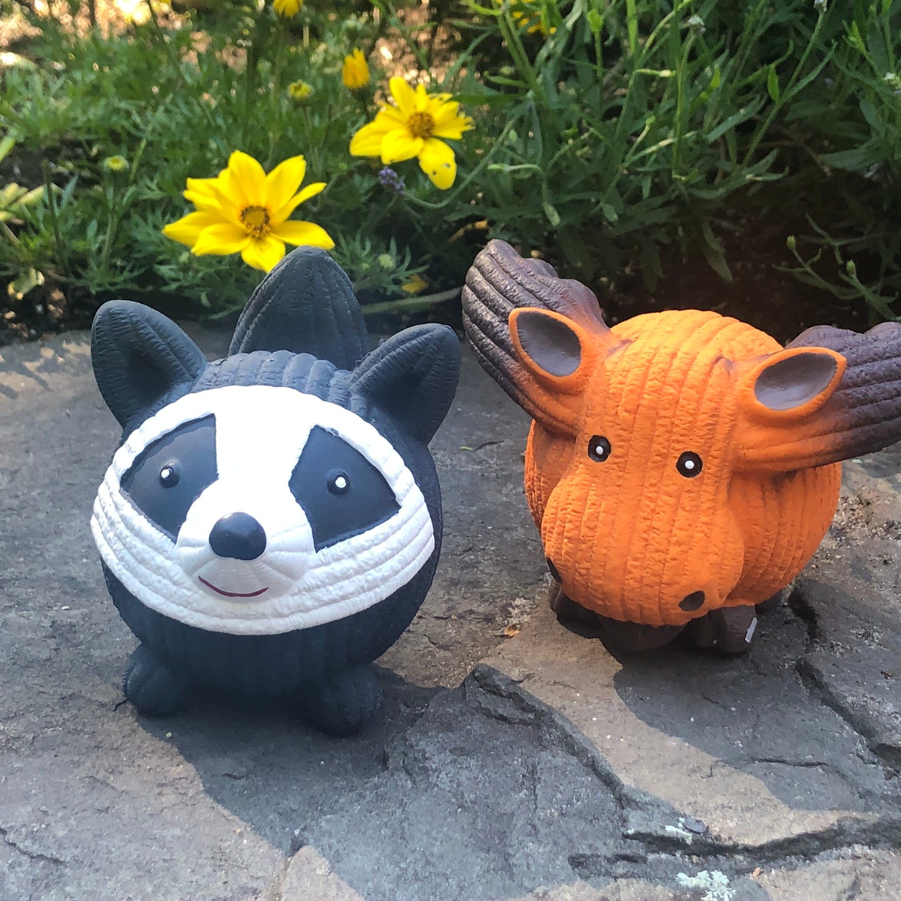 Lifestyle image of Reggie the Raccoon sitting on a rock slab next to Morris the Moose. In the background are green bushes and yellow flowers.
