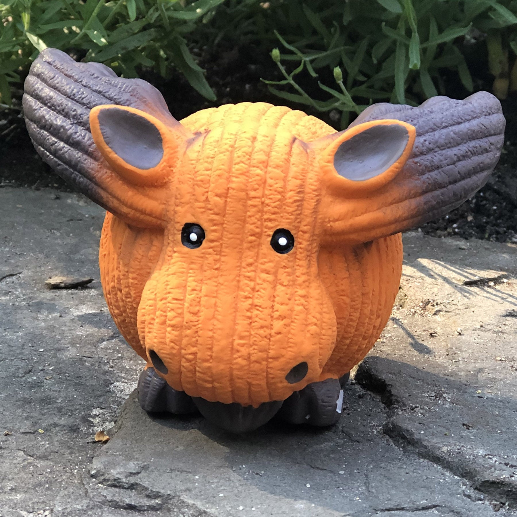 Lifestyle image of Morris the Moose sitting on a rock slab, with green bushes in the background.