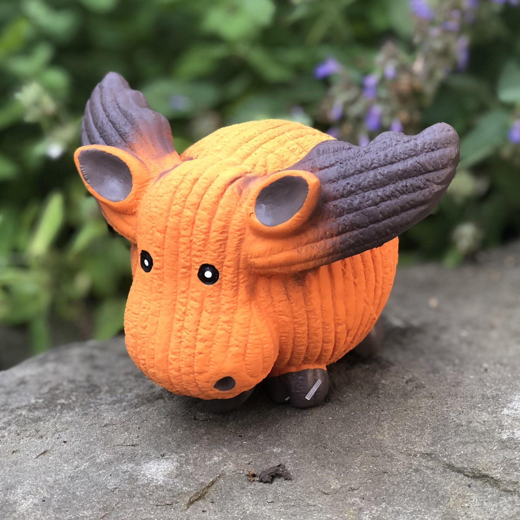 Lifestyle image of Morris the Moose taken from the right side. Morris is sitting on a rock slab with green bushes in the background.