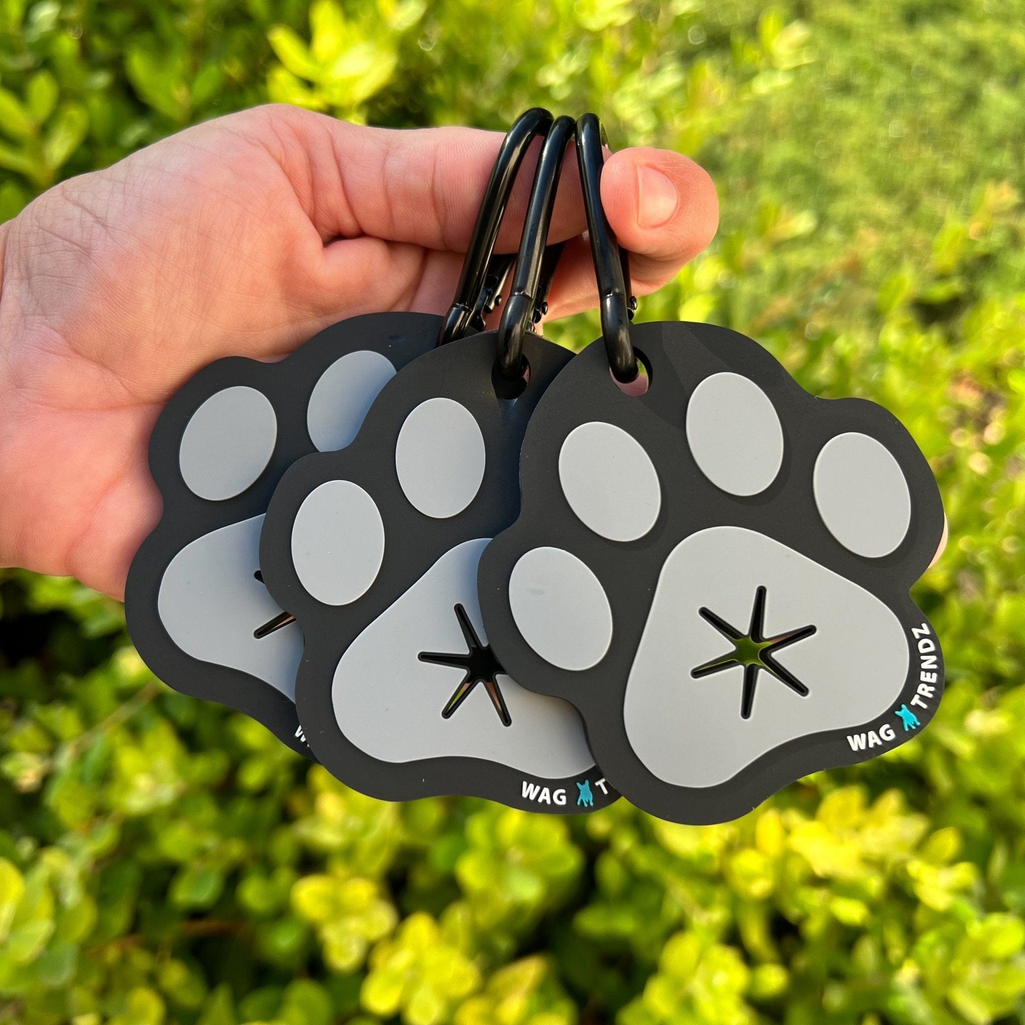Poop Buddy - black and gray resin dog paw - set of 3 held by a hand - outdoors with greenery in the background - Wag Trendz