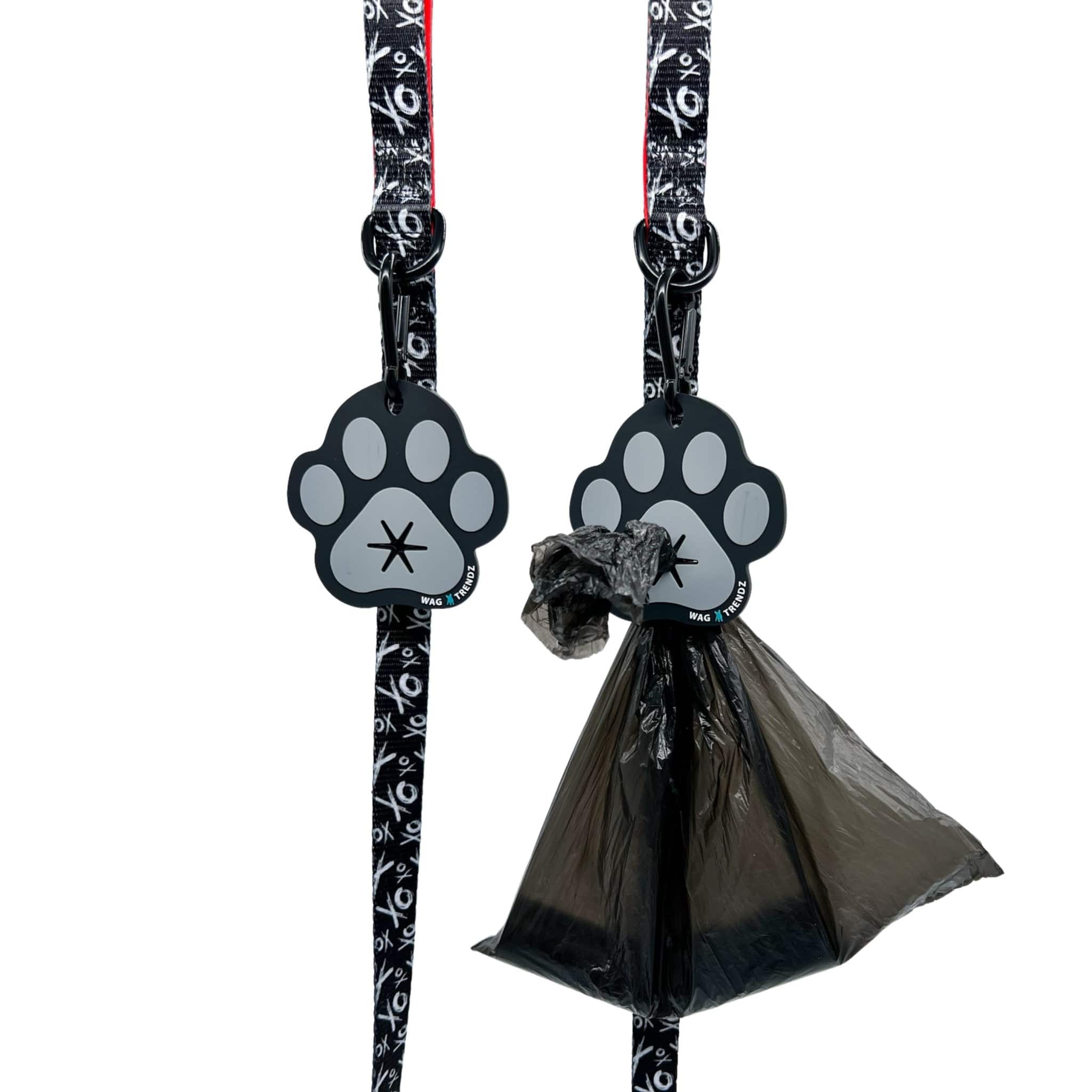 Poop Buddy - black and gray resin dog paw - two hanging on two black & white dog leashes XO with red accents and black plastic bag carrying poop pushed through hole on one - against white background - Wag Trendz