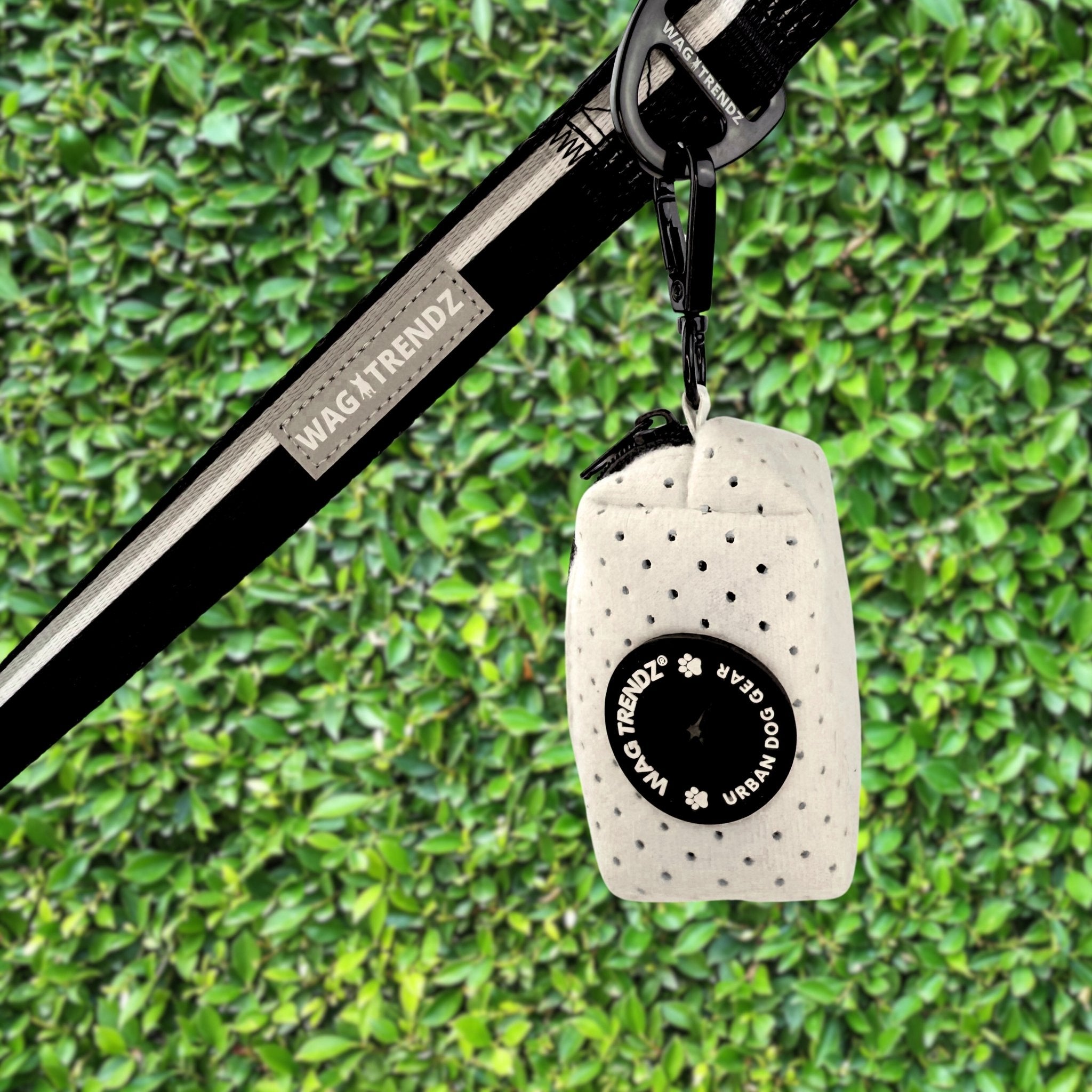 NeoCool™ Sport Poop Bag Holder in White with black logo dispenser attached to a leash against a green hedge background