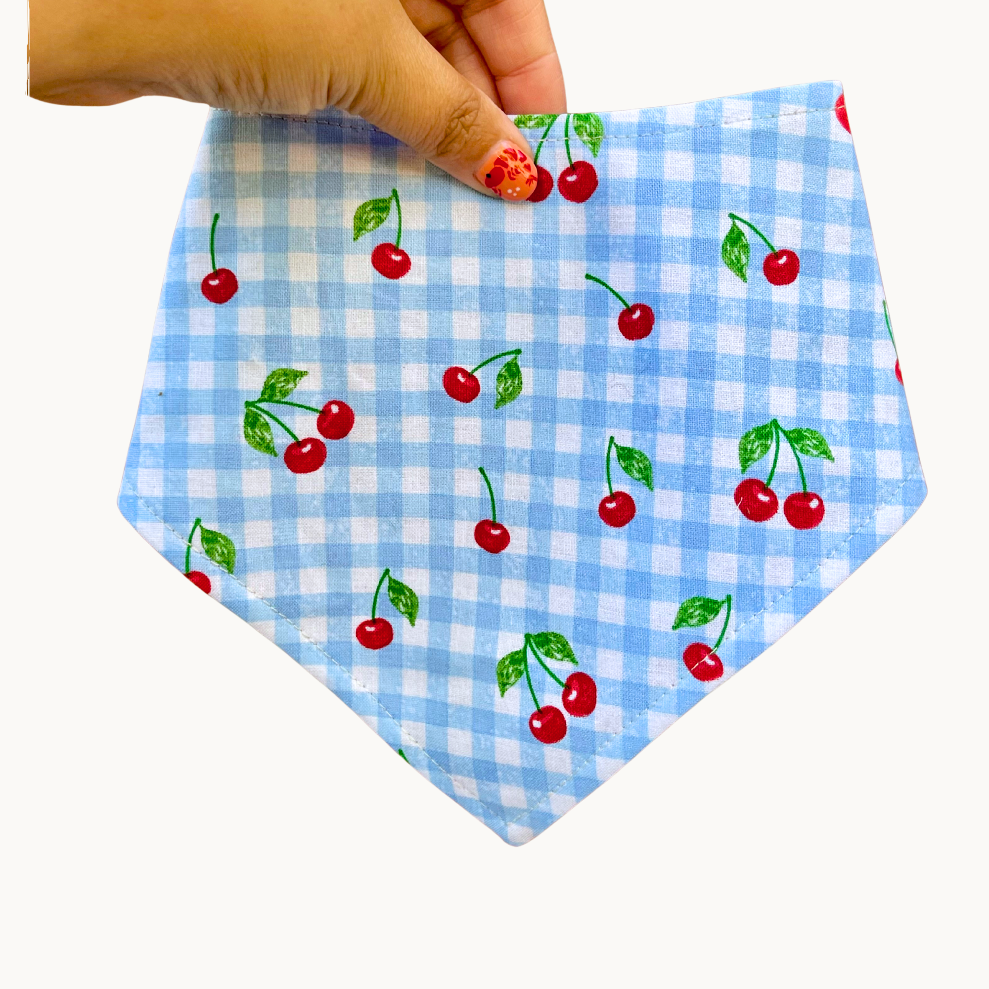 Light blue and white gingham with red cherries handmade dog bandana held in hand on a cream background.